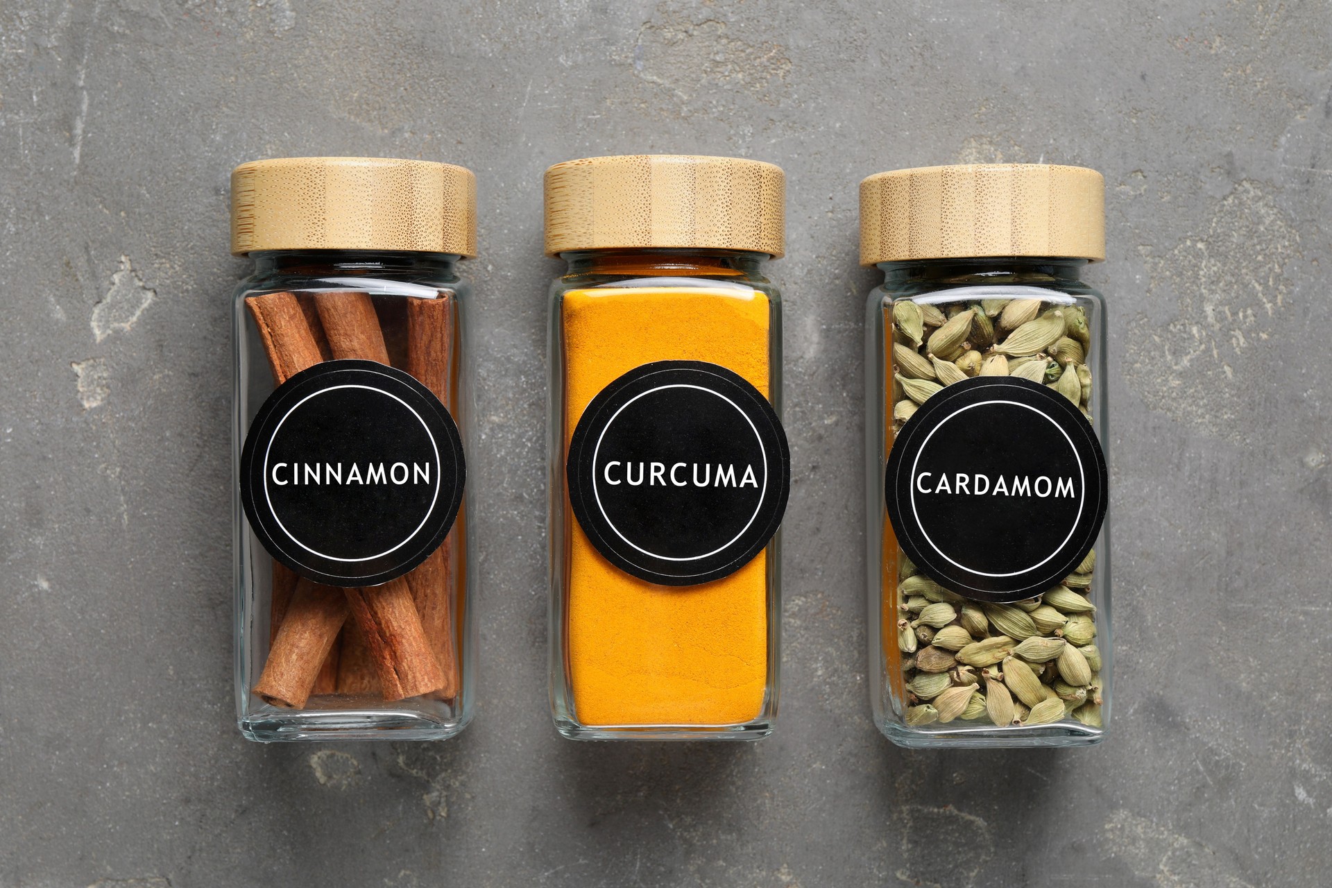 Labelled jars with spices on grey table, flat lay. Cinnamon, curcuma and cardamom